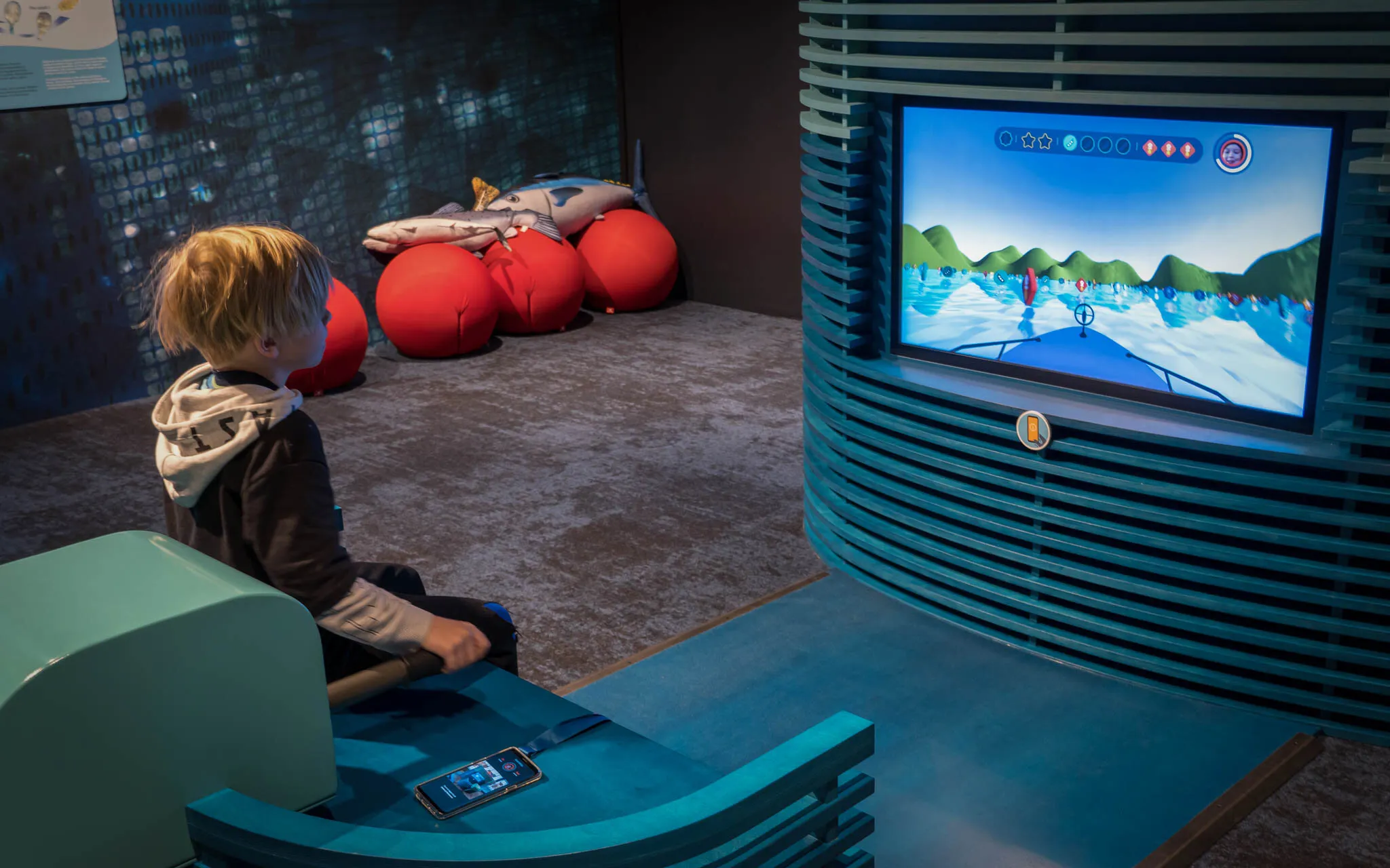 Kid sitting on a blue boat shaped chair holding a boat motor model with a screen in front showing a first person view of driving a boat.