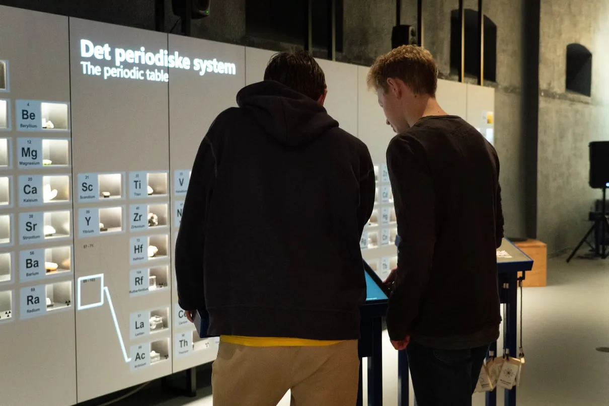 Two people using a touch screen stand in front of a periodic system wall.