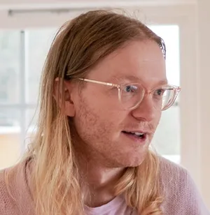 Person with long blond hair, light stubbel, light skin, pink-transparent glasses looking intently to the right