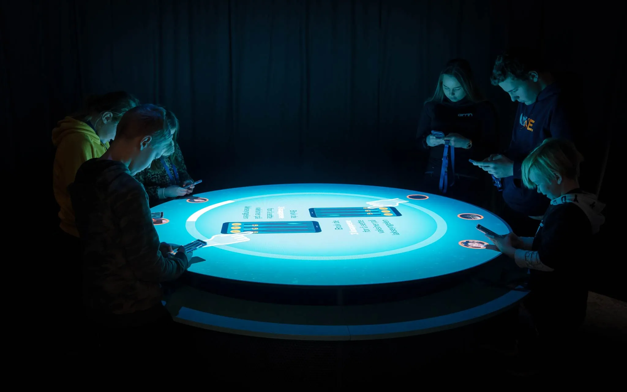 A bunch of people standing around a round table screen, all looking at their phones. The screen gives instructions for what to do on their phones.